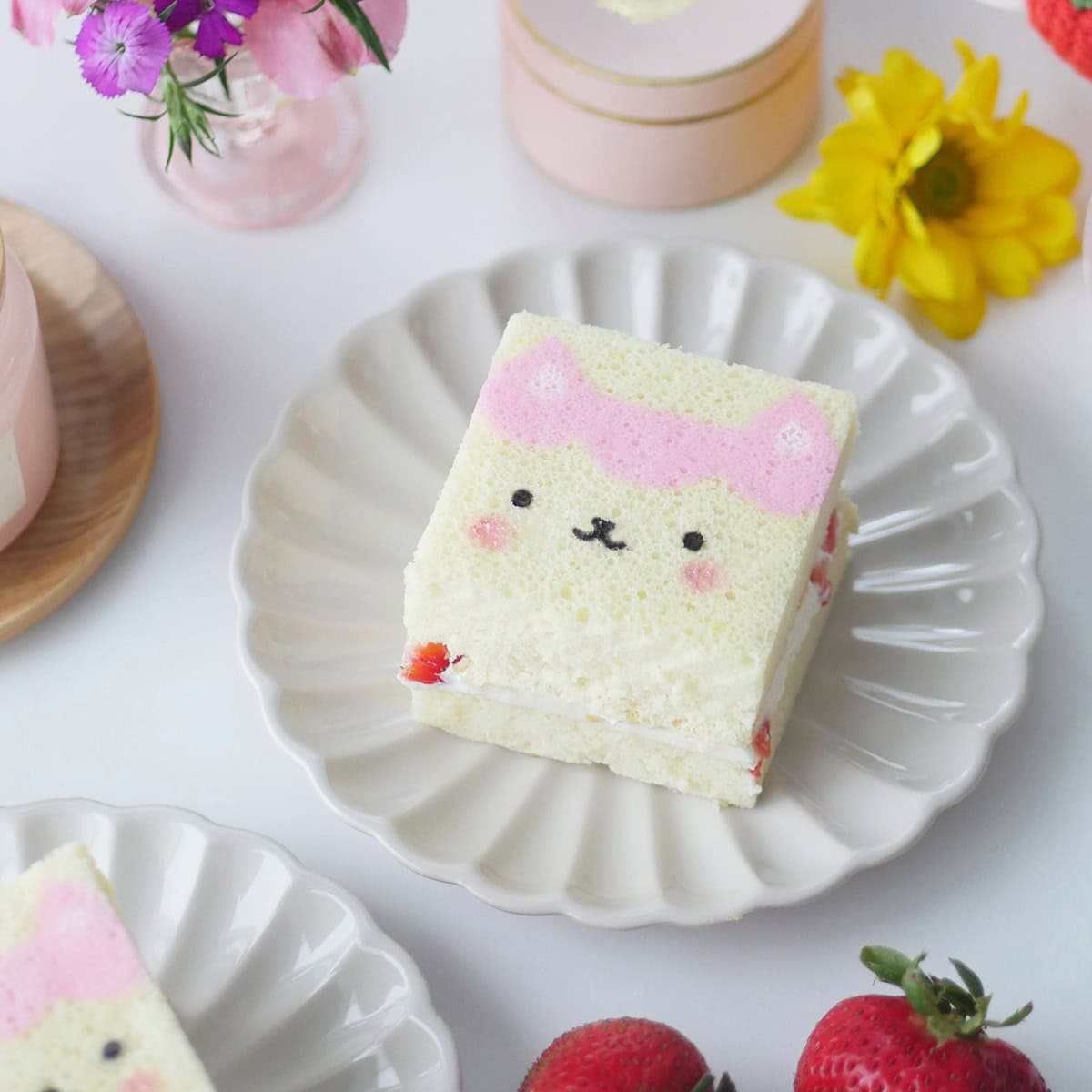 A slice of printed cake of a Gabby Dollhouse character sits on a scalloped plate surrounded by flowers.