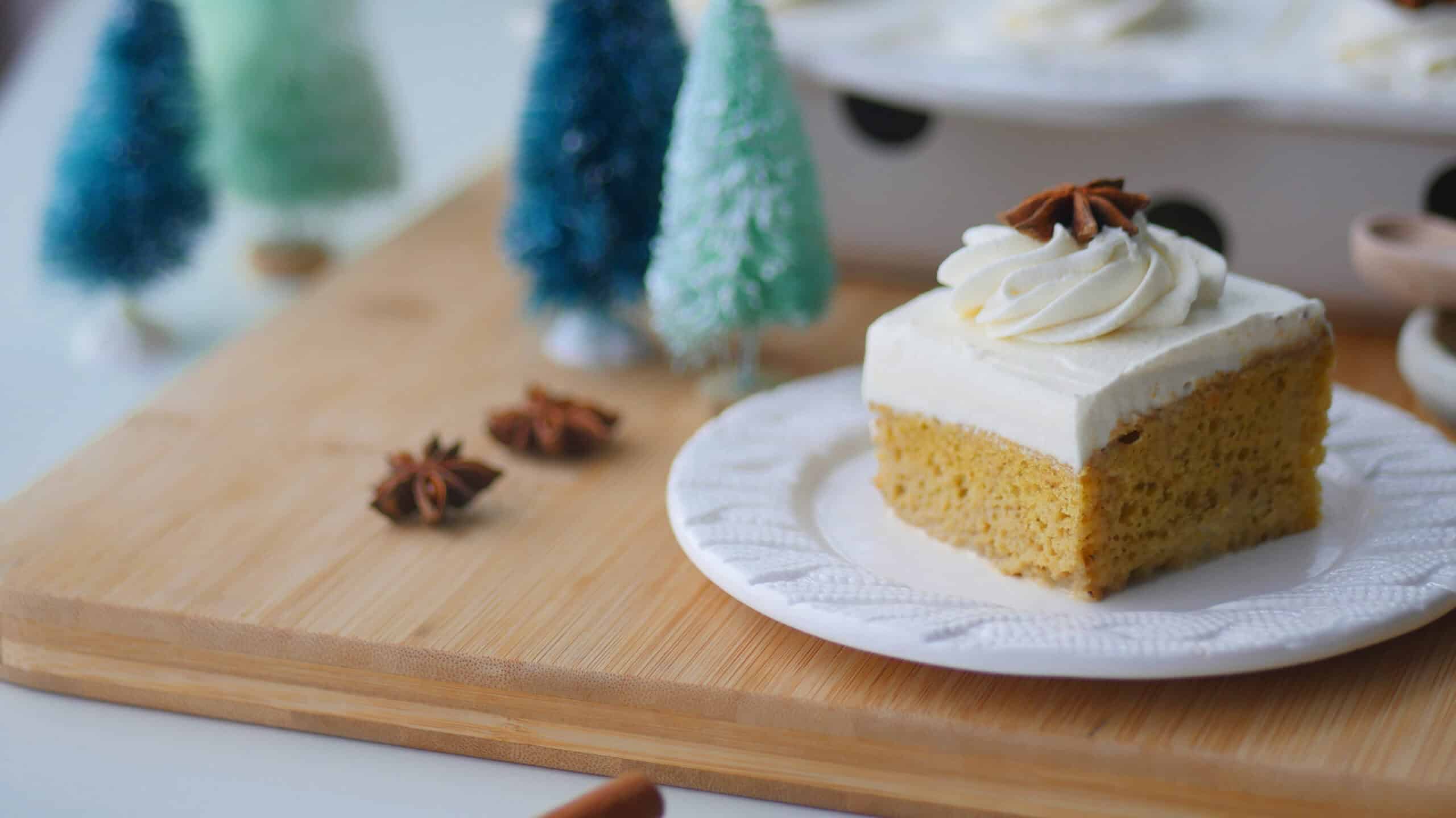 A landscape oriented photo of a slice of gingerbread tres leches cake on a plate with the whole pan in the background along with some bottle brush Christmas trees.